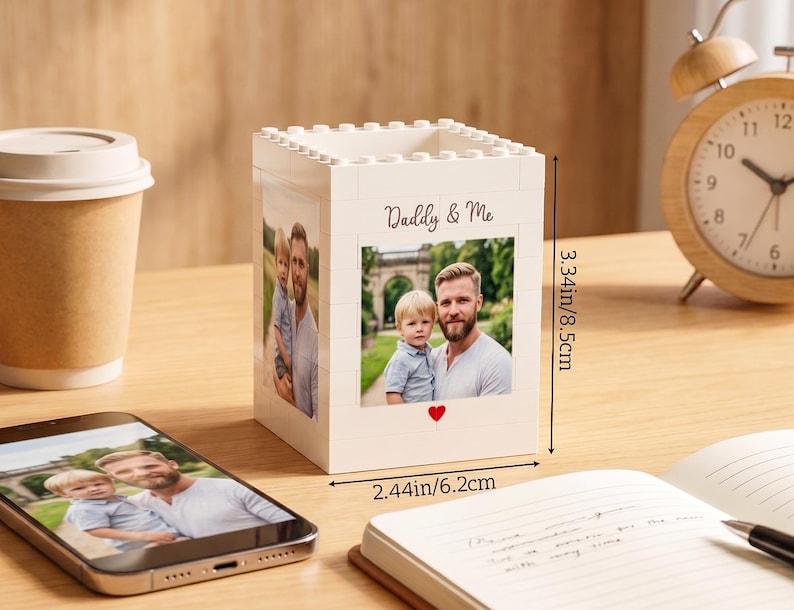 以下が含まれることがあります： 「Daddy & Me」の文字と父と子の写真が入った、白いブロック型のフォトホルダー。ホルダーの高さは8.5cm、幅は6.2cmです。写真の下には赤いハートがあります。