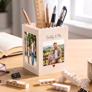 May include: A cream-colored desk organizer made of interlocking blocks, featuring two framed photos and the text "Daddy & Me." The organizer holds pencils, a ruler, and pens. A coffee cup, open book, and glasses are on the wooden desk.
