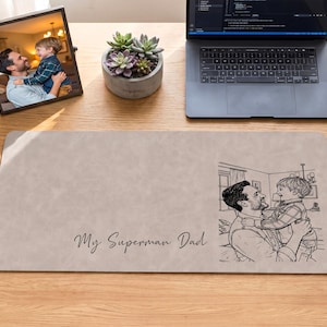 May include: A beige desk mat with the text "My Superman Dad" and a sketch of a father and child. A framed photo and a succulent plant are also on the wooden desk.