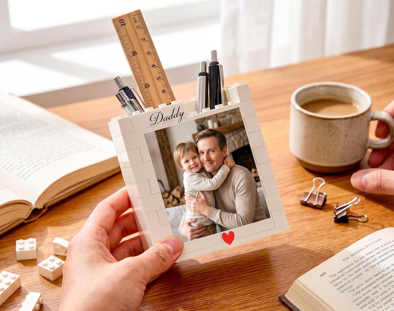 以下が含まれることがあります： 「Daddy」と赤いハートのラベルが付いた、男性と子供の写真が入った白いレンガ風のペン立て。木製の定規とペンがホルダーに入っています。コーヒーカップと開いた本が木製のテーブルの上にあります。