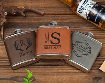 May include: Three personalized leather-wrapped hip flasks with engraved designs. The flasks are brown, gray, and tan. The first flask has a stag head design with the letter R and the name Ricard. The second flask has the name Scott and the letter S with the words Best Man. The third flask has the name Ethan and the date 8.15.2025 with the words Groomman.