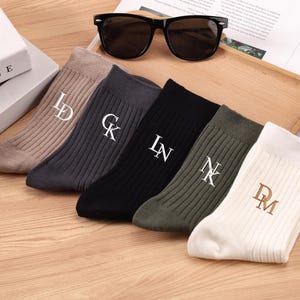 Puede incluir: Seis pares de calcetines acanalados en varios colores, beige, gris, negro y verde oliva, cada uno bordado con iniciales blancas o marrones. Unas gafas de sol negras descansan sobre una bandeja de madera.