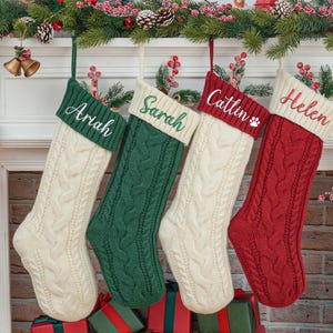 Puede incluir: Cuatro calcetines de Navidad de punto personalizados colgados en una repisa de chimenea blanca. Los calcetines son blancos, verdes, rojos y crema con nombres bordados en blanco: Ariah, Sarah, Caitlin y Helen.