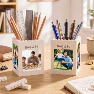 以下が含まれることがあります： 家族写真と「Daddy & Me」の文字が入った、クリーム色のレンガ風ペン立てが2つ。1つは鉛筆と定規、もう1つはペンが入っています。木製の机の上にはコーヒーカップ、ノート、文房具が置かれています。