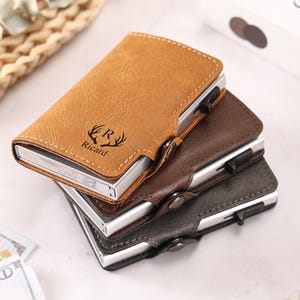 May include: Three leather cardholders in tan, brown, and grey stacked. The top cardholder has the name "Ricard" and a deer antler design. Each cardholder has a metal frame and a button closure.