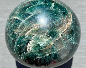May include: A polished, spherical gemstone with a predominantly green hue, featuring intricate patterns and subtle variations in color. The surface reflects light, creating highlights and shadows. The sphere's diameter appears to be approximately 3 inches.