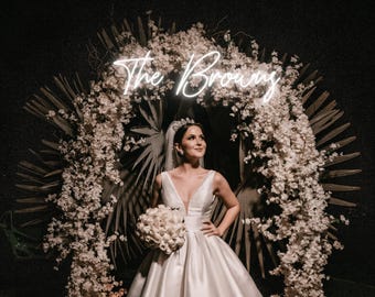 May include: A bride in a white satin wedding dress stands in front of a floral arch decorated with white flowers and greenery. The arch has a neon sign that reads "The Browning".