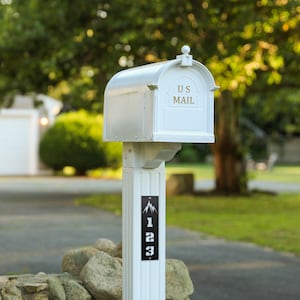 Può includere: Una cassetta postale bianca con la parte superiore curva e le parole "US MAIL" sulla parte anteriore. La cassetta postale è montata su un palo bianco con una targa con l'indirizzo nero che recita "123". Lo sfondo è un paesaggio verde sfocato.