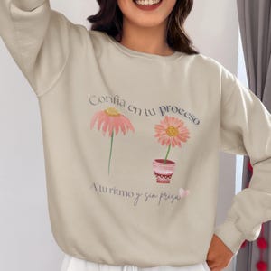 Puede incluir: Sudadera beige con diseño floral y texto en español. El diseño presenta dos flores rosas, una con tallo y otra en maceta. El texto dice "Confía en tu proceso" y "A tu ritmo y sin prisa".