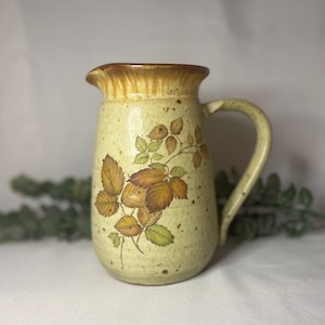 May include: A ceramic pitcher with a brown rim and a beige body. The pitcher has a floral design with brown and green leaves.
