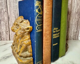 May include: A pair of wooden bookends shaped like a group of three dogs. The bookends are holding up four books, including "Tom Cringle's Log" and "Smiley People" by John le Carre.