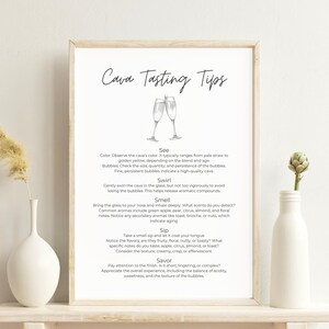 Op de afbeelding: Een ingelijste print met de tekst "Cava Tasting Tips" en illustraties van champagneglazen. De print wordt weergegeven op een witte plank met een witte vaas en een gedroogde plant links en een witte fles met een groene plant rechts.