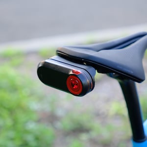 Puede incluir: Una luz de bicicleta negra con una lente roja unida a un sillín de bicicleta negro.