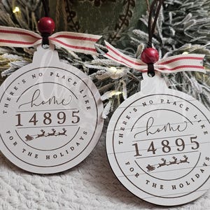 May include: Two round white wooden ornaments featuring the text "THERE'S NO PLACE LIKE home 14895 FOR THE HOLIDAYS" in dark brown lettering. Each ornament includes a small red bead, a brown string for hanging, and a red and white striped ribbon.