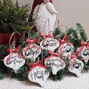 May include: White Christmas ornaments with black script and red ribbon bows. The ornaments have words like "Believe", "Grace", and "Joy". They are arranged on a bed of greenery, with a red bucket and a decorative figure in the background.