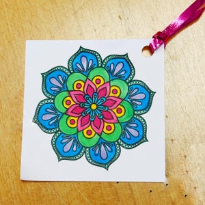 May include: A colorful mandala design on a white square tag with a hole punched at the top. The mandala features a symmetrical floral pattern in shades of pink, blue, green, and yellow. A pink ribbon is attached.