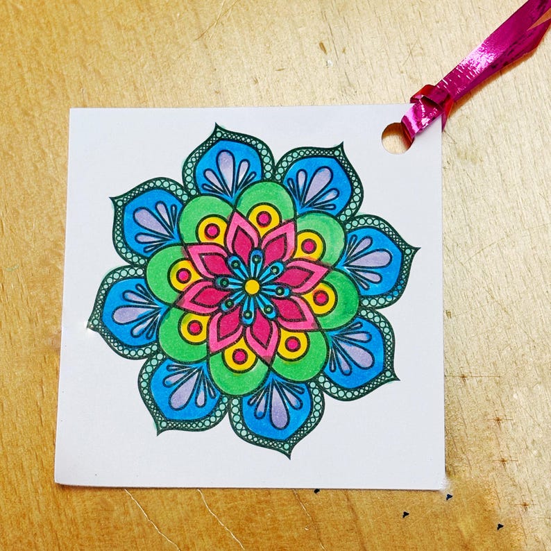 May include: A colorful mandala design on a white square tag with a hole punched at the top. The mandala features a symmetrical floral pattern in shades of pink, blue, green, and yellow. A pink ribbon is attached.
