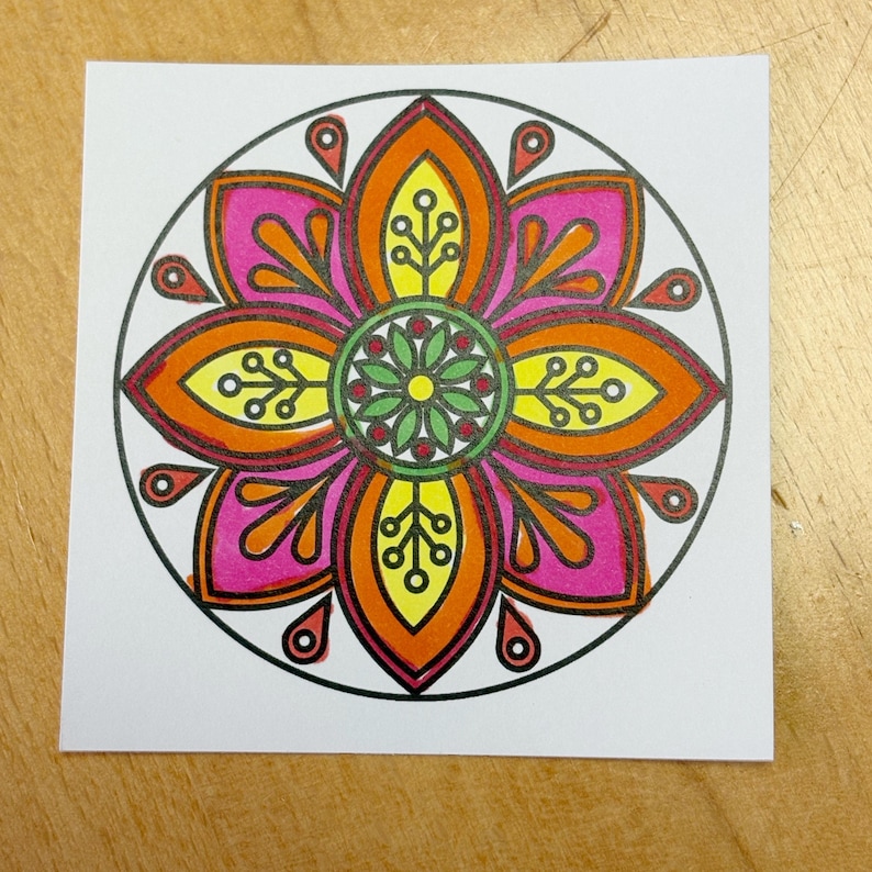 May include: A colorful mandala design on a square white paper. The mandala features a central flower with pink, orange, and yellow petals, outlined in black. The design includes small teardrop shapes and a circular border.