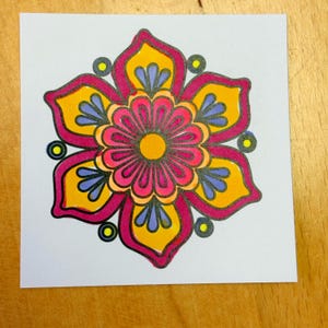 May include: A colorful floral design on a white square. The flower features a central pink bloom with a yellow center, surrounded by petals in yellow, pink, and blue. The design is outlined in black, with small yellow circles around the petals.