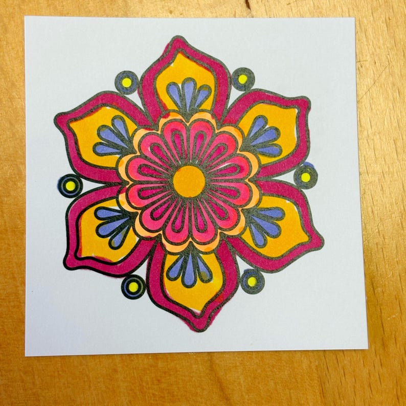 May include: A colorful floral design on a white square. The flower features a central pink bloom with a yellow center, surrounded by petals in yellow, pink, and blue. The design is outlined in black, with small yellow circles around the petals.