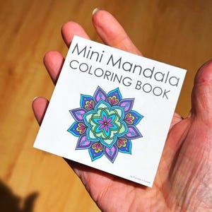 May include: A mini mandala coloring book held in a hand. The cover features the text "Mini Mandala COLORING BOOK" above a colorful mandala design with blue, purple, pink, and green hues. The book is small and square.