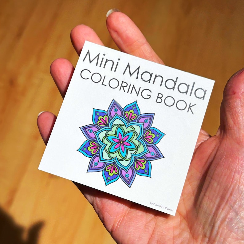 May include: A mini mandala coloring book held in a hand. The cover features the text "Mini Mandala COLORING BOOK" above a colorful mandala design with blue, purple, pink, and green hues. The book is small and square.