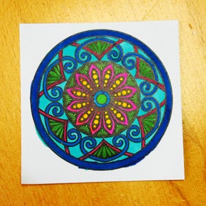 May include: A colorful mandala design on a square white paper. The mandala features a central flower with pink petals and yellow dots, surrounded by teal, green, and blue geometric patterns. The design is enclosed within a dark blue border.