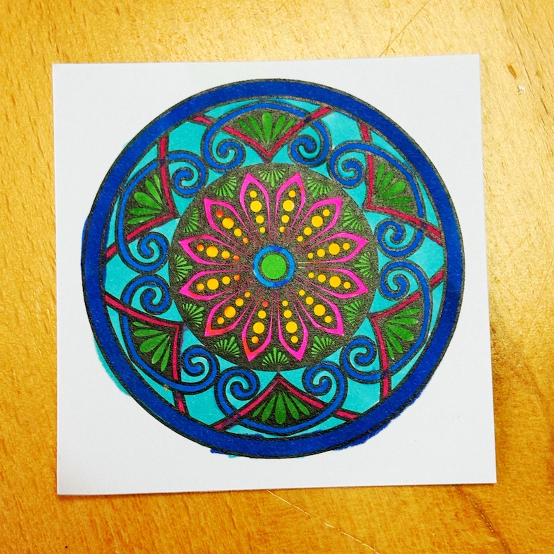 May include: A colorful mandala design on a square white paper. The mandala features a central flower with pink petals and yellow dots, surrounded by teal, green, and blue geometric patterns. The design is enclosed within a dark blue border.