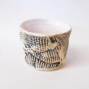 May include: A small, white ceramic cup with a textured, black and white design. The cup has a slightly rounded shape and a wide, open top.