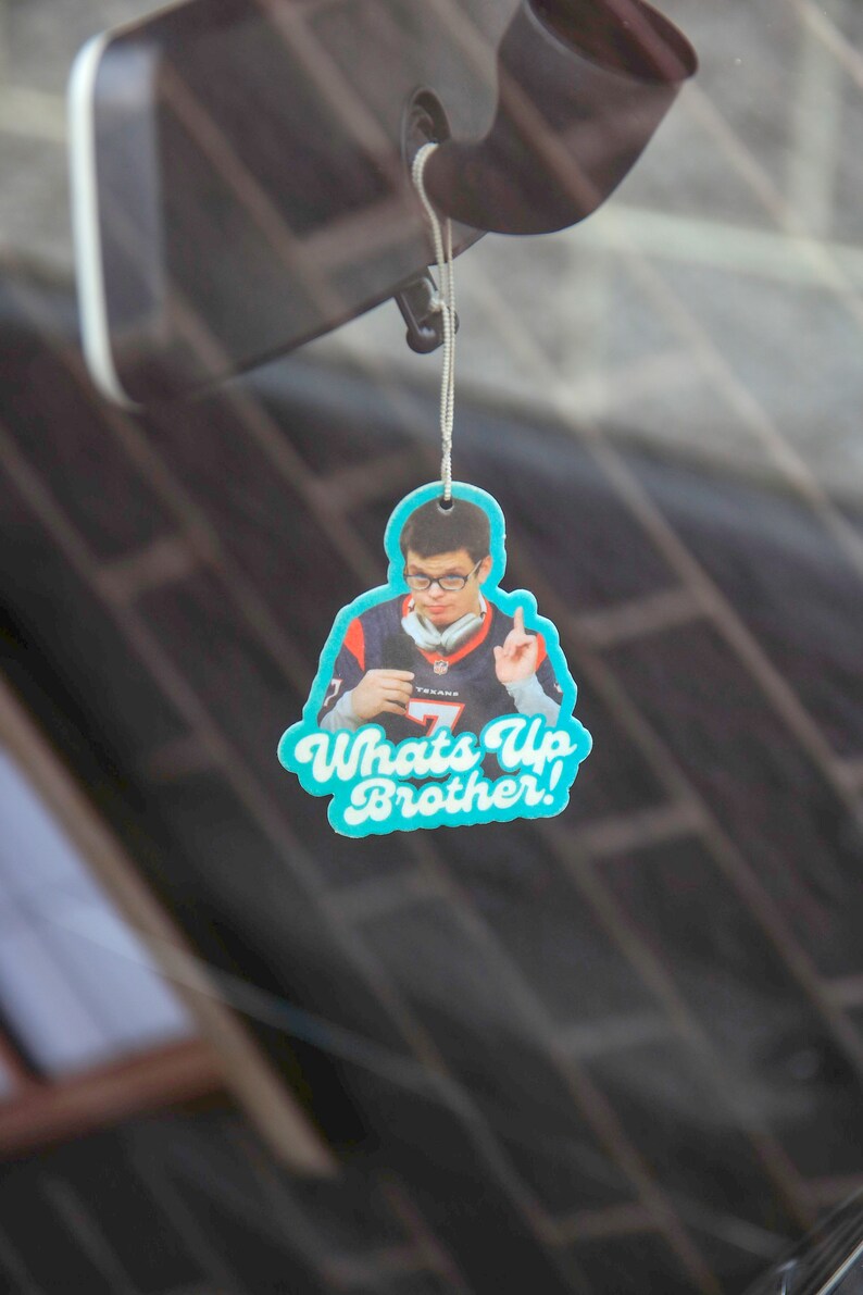 May include: A blue and white car air freshener with a photo of a person wearing a jersey and the text "Whats Up Brother!"
