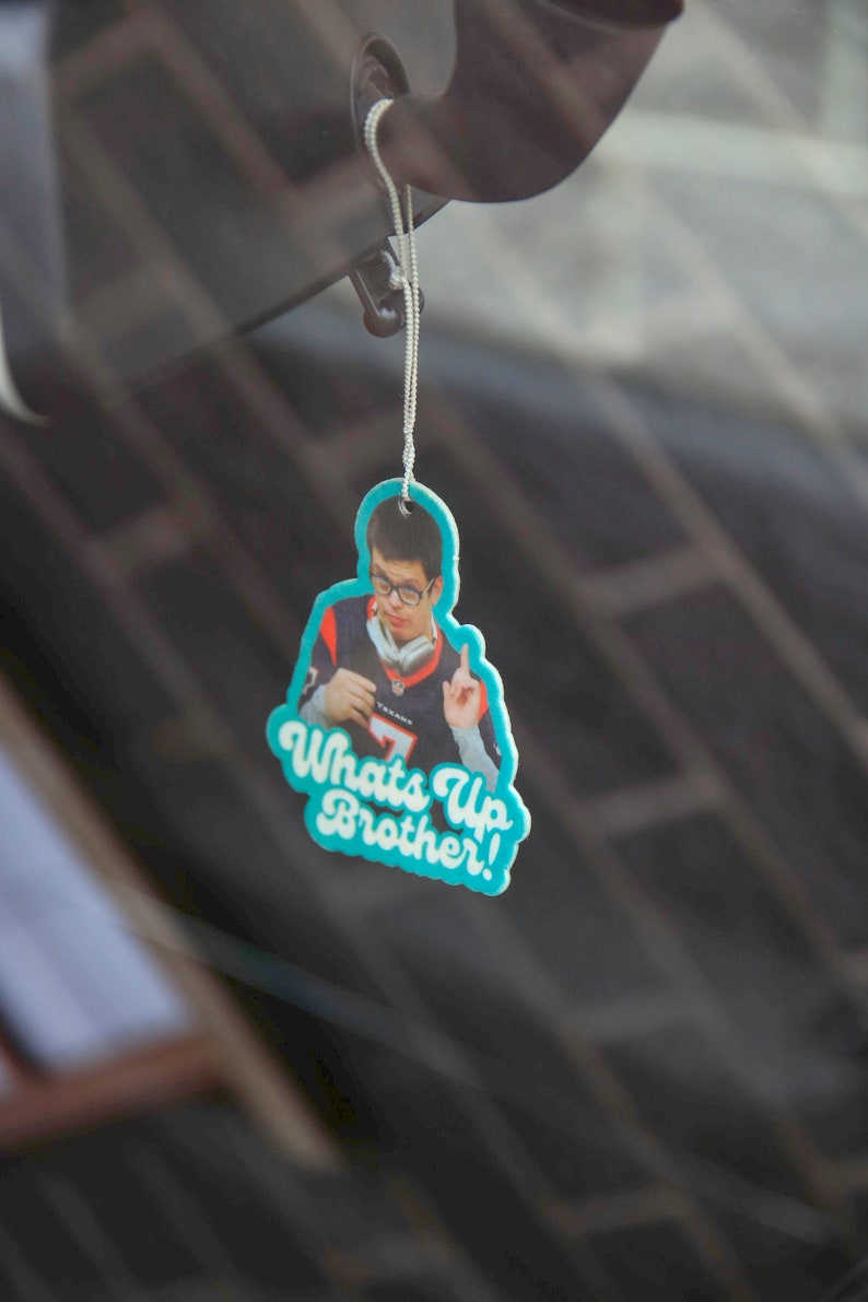 May include: A blue and white car air freshener with a photo of a man wearing a jersey and the text "Whats Up Brother!"