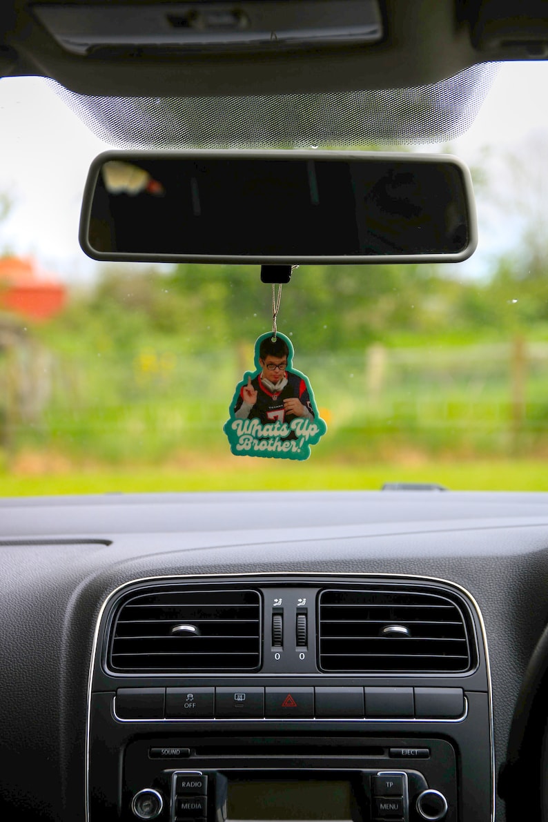 May include: A green car air freshener with a photo of a person wearing a jersey and the text "What's Up Brother!" hanging from the rearview mirror of a car.