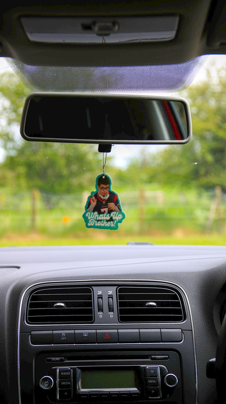 May include: A teal car air freshener with a photo of a man wearing a blue and white jersey. The text on the air freshener reads "Whats Up Brother!"