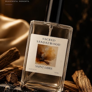May include: A clear glass perfume bottle with a black cap, labeled "SACRED SANDALWOOD" and "SAINT LIBRE." The bottle is 100 ml and made in the USA. The image includes wood pieces and the text "TIMELESS SCENTS. MEANINGFUL MOMENTS."