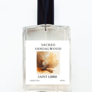May include: A clear glass perfume bottle with a black cap. The label reads "Sacred Sandalwood" and "Saint Libre." The label features an illustration of incense and sticks. The bottle contains 100 mL of product and is made in the USA.