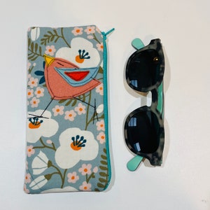 May include: A blue floral sunglasses case with a bird applique and a pair of sunglasses. The case has a teal zipper and a bird applique with a pink body, blue wings, and a yellow beak. The sunglasses have a gray and black speckled frame and teal arms.