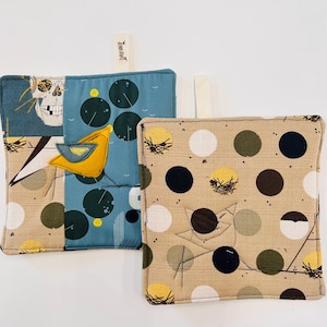 Puede incluir: Dos agarraderas cuadradas de tela. Una presenta una calavera, nenúfares y un bote amarillo sobre un fondo azul. La otra tiene un fondo beige con lunares grandes en negro, blanco, amarillo y marrón. Cada una tiene un lazo blanco.