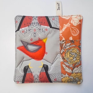 May include: Square quilted potholder with a red bird applique and a patchwork design. The bird has a yellow beak and gray accents. The patchwork includes orange floral and geometric patterns. A fabric loop is attached for hanging.