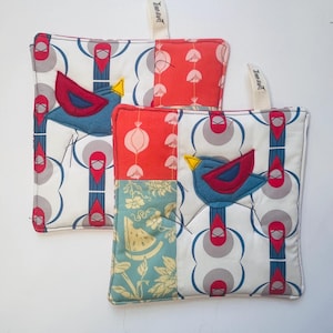 May include: Two square quilted pot holders with a patchwork design. One side features a blue bird applique with red wings and a yellow beak. The other side has a mix of red and teal patterned fabrics. Each pot holder has a fabric loop for hanging.