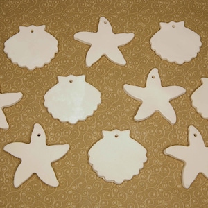 May include: A collection of ten white ceramic ornaments in the shapes of starfish and scallop shells. Each ornament has a small hole at the top for hanging. The ornaments are arranged on a patterned, golden-brown surface.