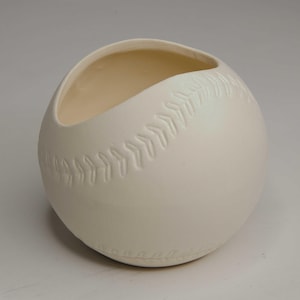 May include: A baseball-shaped ceramic vase in off-white. The vase has a curved opening and embossed stitching details mimicking a baseball. The interior is a lighter cream color. A unique decorative piece for sports enthusiasts.