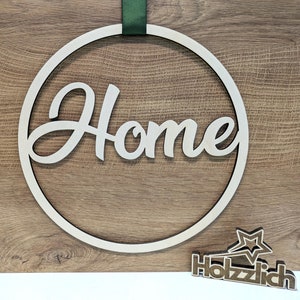 Könnte beinhalten: Ein Holz-Kreis mit dem Wort "Home" in kursiver Schrift. Der Kreis hängt an einem grünen Band. Das Wort "Holzzlich" steht auf einem kleinen Holzstern.