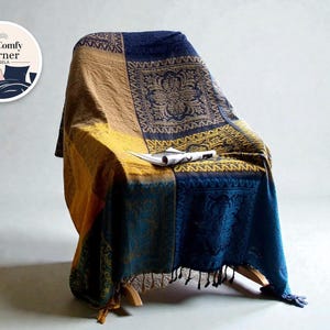 May include: A patterned throw blanket in navy blue, mustard yellow, and tan, draped over a wooden chair. The blanket has detailed designs. A newspaper rests on the blanket. The Comfy Corner by Eysela logo is in the upper left corner.