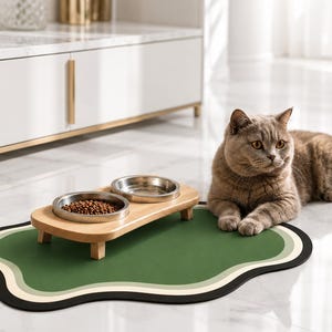 May include: A cat food and water bowl set on a green and black mat. The wooden stand holds two stainless steel bowls, one filled with kibble. A gray cat is lying next to the mat on a white tile floor.