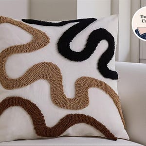 Op de afbeelding: Vierkant wit decoratief kussen met een textuurontwerp. Het kussen heeft een patroon van vloeiende, abstracte vormen in zwart, beige en bruin. Het kussen ligt op een witte bank. Het logo The Comfy Corner by SSEL staat rechtsboven.