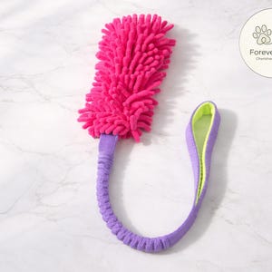 Könnte beinhalten: Ein leuchtend rosa Hundespielzeug mit einem strukturierten Kopf und einem lila Griff. Der Griff hat eine limettengrüne Innenseite und ist mit lila Stoff am Kopf befestigt. Das Spielzeug ist für Haustiere konzipiert.