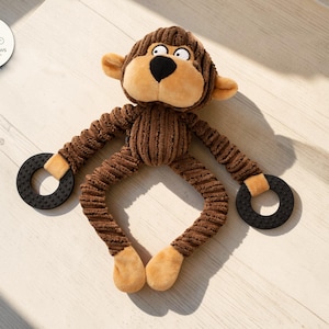 May include: Brown plush monkey dog toy with a corduroy body, tan accents, and black rubber rings on the arms. The monkey has a friendly expression with large eyes and a black nose. The toy is designed for dogs.
