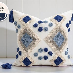 Könnte beinhalten: Ein weißes Zierkissen mit geometrischem Design. Das Kissen hat ein Rautenmuster in Blau- und Beigetönen mit getufteten Akzenten und Quasten. Das Kissen misst etwa 46 cm im Quadrat.