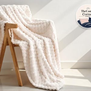 May include: A cream-coloured, textured blanket draped over a wooden chair. The blanket has a plush, bubble-like pattern. The chair is made of light-coloured wood. The Comfy Corner logo is visible in the upper right corner.