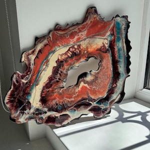 May include: A unique, free-form wall art piece with a central opening. The artwork features a layered design with shades of red, orange, cream, and teal, framed by a black border. The piece is displayed on a white wall.